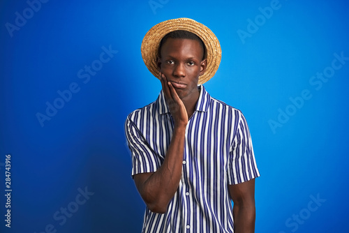 African american man wearing striped shirt and summer hat over isolated blue background thinking looking tired and bored with depression problems with crossed arms.