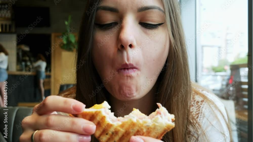 Close-up portrait of young hungry woman with pleasure eats toast in a cafe, fast food restaurant Close-up portrait of young hungry woman with pleasure eats toast in a cafe, fast food restaurant
