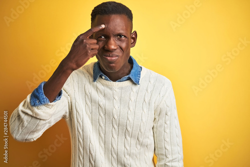 Obraz na plátně African american man wearing denim shirt and white sweater over isolated yellow background pointing unhappy to pimple on forehead, ugly infection of blackhead