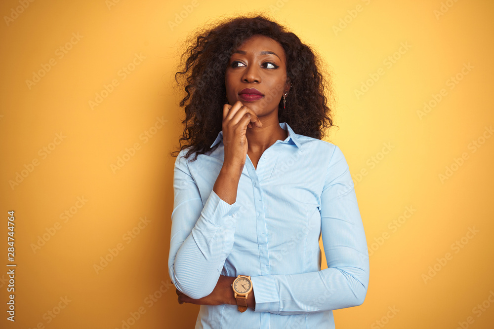 African american businesswoman wearing elegant shirt over isolated yellow background with hand on chin thinking about question, pensive expression. Smiling with thoughtful face. Doubt concept. African american businesswoman wearing elegant shirt over isolated yellow background with hand on chin thinking about question, pensive expression. Smiling with thoughtful face. Doubt concept.