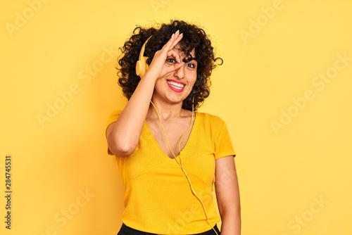 Arab woman with curly hair listening to music using headphones over isolated yellow background doing ok gesture with hand smiling, eye looking through fingers with happy face.