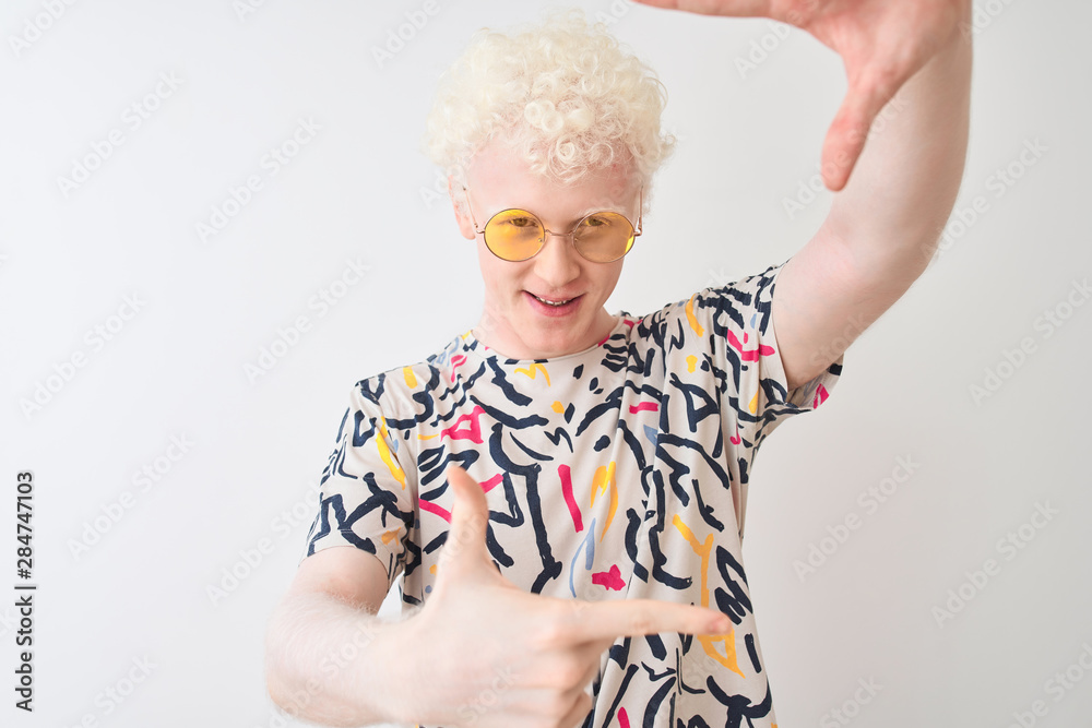 Young albino blond man wearing colorful t-shirt and sunglasses over isolated red background smiling making frame with hands and fingers with happy face. Creativity and photography concept.