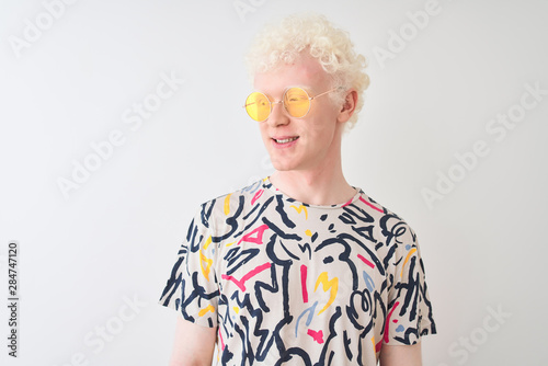 Young albino blond man wearing colorful t-shirt and sunglasses over isolated red background looking away to side with smile on face, natural expression. Laughing confident.