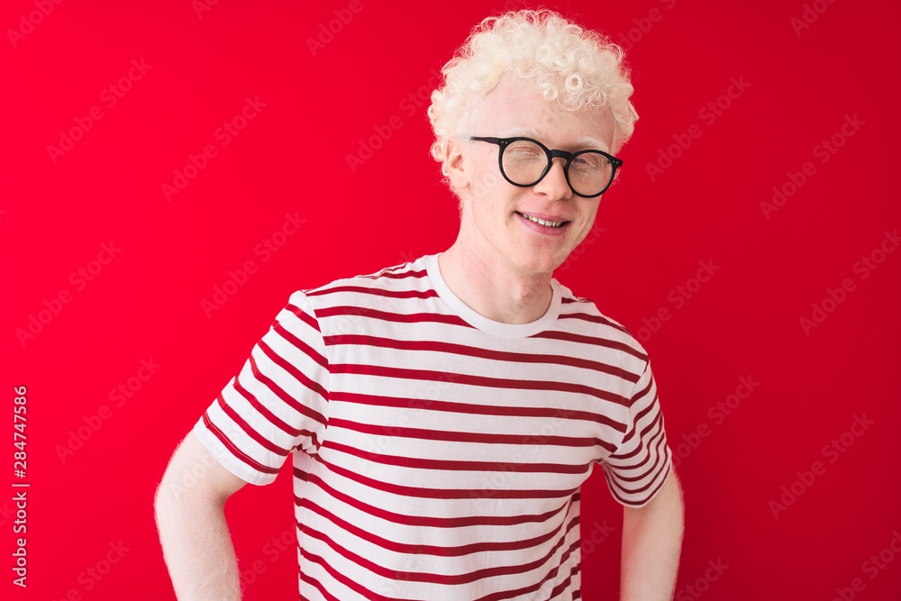 © Krakenimages.com - Young albino blond man wearing striped t-shirt and glasses over isolated red background winking looking at the camera with sexy expression, cheerful and happy face.