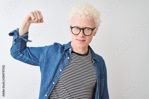 Young albino blond man wearing denim shirt and glasses over isolated white background Strong person showing arm muscle, confident and proud of power