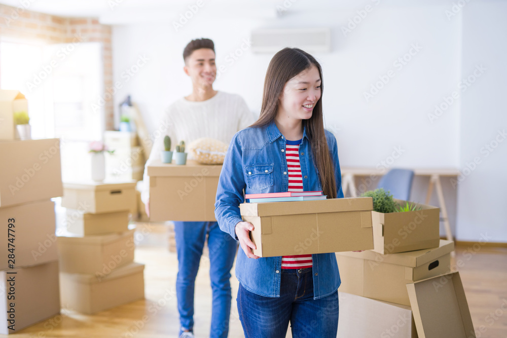 Beautiful young asian couple looking happy holding cardboard boxes, smiling excited moving to a new home