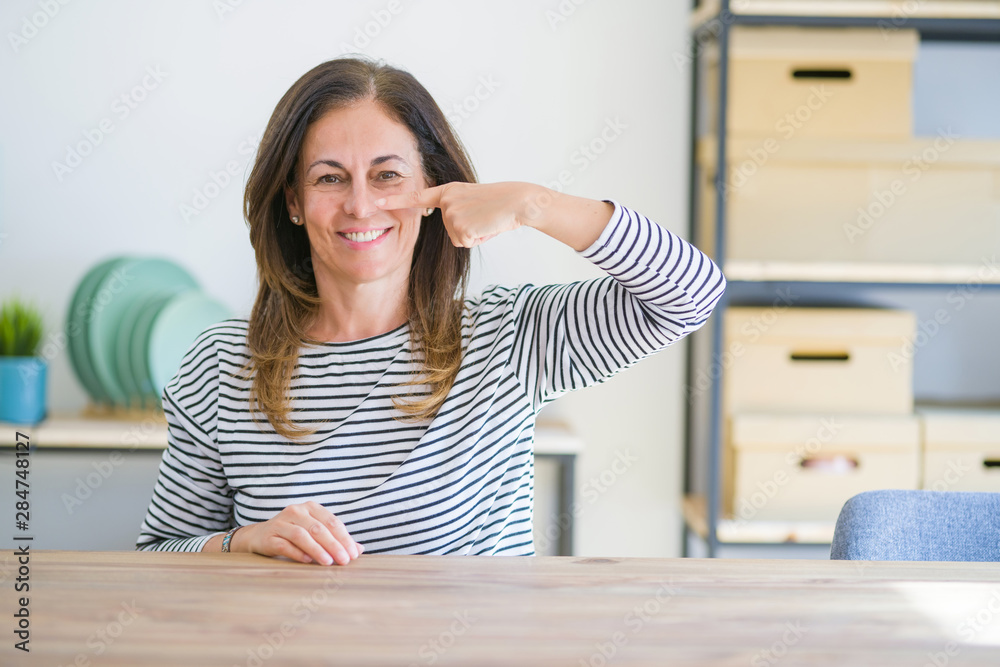 Middle age senior woman sitting at the table at home Pointing with hand finger to face and nose, smiling cheerful. Beauty concept Middle age senior woman sitting at the table at home Pointing with hand finger to face and nose, smiling cheerful. Beauty concept
