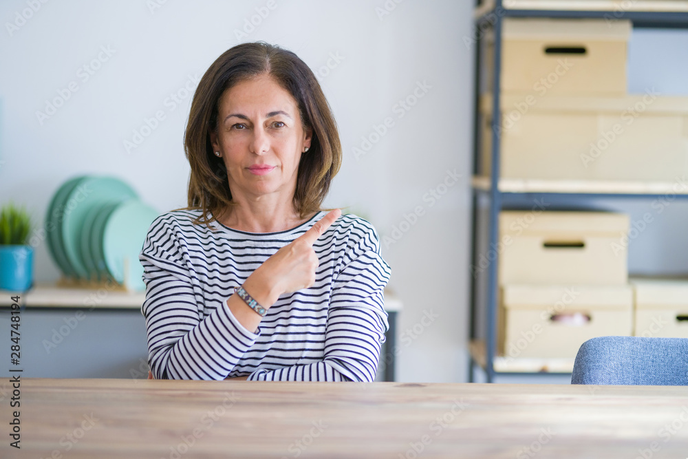Middle age senior woman sitting at the table at home Pointing with hand finger to the side showing advertisement, serious and calm face Middle age senior woman sitting at the table at home Pointing with hand finger to the side showing advertisement, serious and calm face