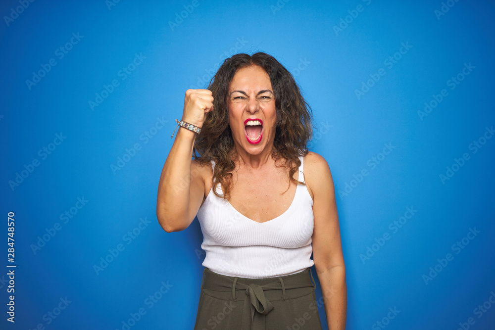 Middle age senior woman with curly hair standing over blue isolated background angry and mad raising fist frustrated and furious while shouting with anger. Rage and aggressive concept. Middle age senior woman with curly hair standing over blue isolated background angry and mad raising fist frustrated and furious while shouting with anger. Rage and aggressive concept.