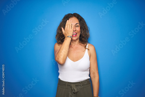 Middle age senior woman with curly hair standing over blue isolated background covering one eye with hand, confident smile on face and surprise emotion.
