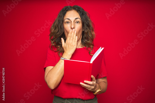 Middle age senior woman reading a book over red isolated background cover mouth with hand shocked with shame for mistake, expression of fear, scared in silence, secret concept