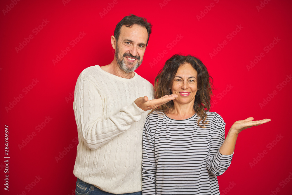 Beautiful middle age couple wearing winter sweater over isolated red background smiling cheerful presenting and pointing with palm of hand looking at the camera. Beautiful middle age couple wearing winter sweater over isolated red background smiling cheerful presenting and pointing with palm of hand looking at the camera.