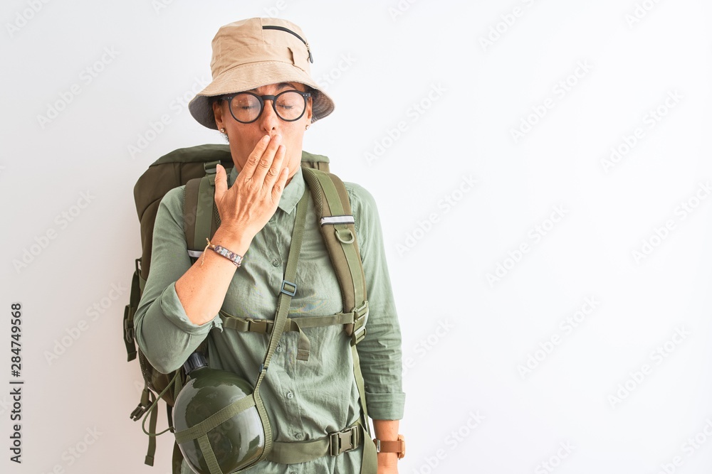 Fototapeta premium Middle age hiker woman wearing backpack canteen hat glasses over isolated white background bored yawning tired covering mouth with hand. Restless and sleepiness.