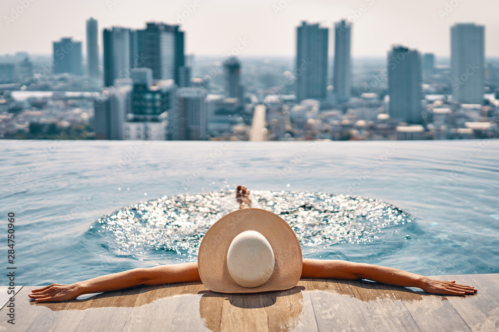 Back view of young woman in hat relaxing in swimming pool on the roof top of hotel - obrazy, fototapety, plakaty 