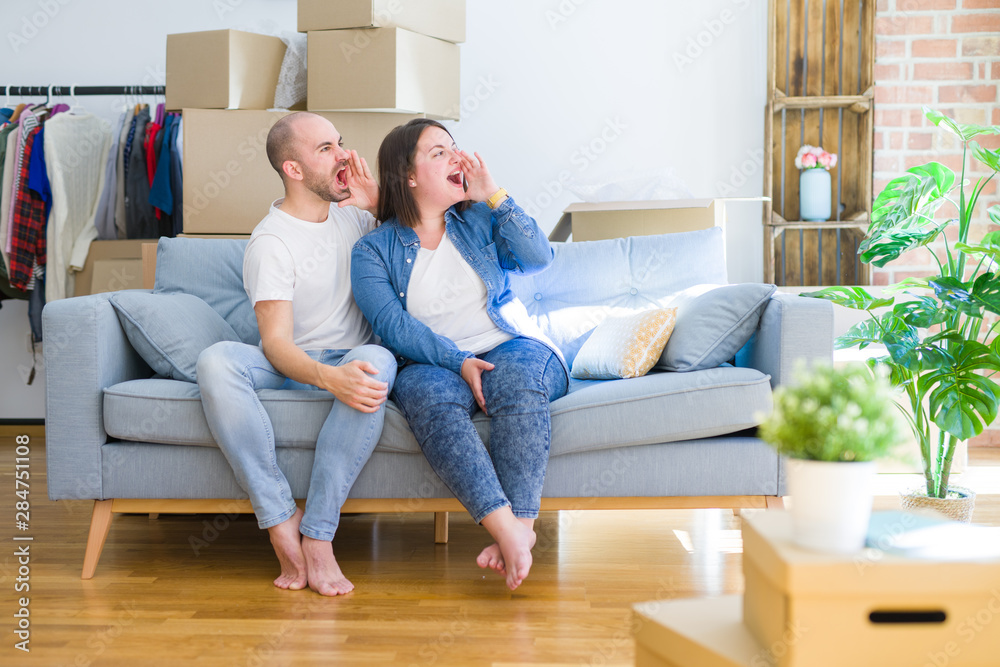 Young couple sitting on the sofa arround cardboard boxes moving to a new house shouting and screaming loud to side with hand on mouth. Communication concept. Young couple sitting on the sofa arround cardboard boxes moving to a new house shouting and screaming loud to side with hand on mouth. Communication concept.