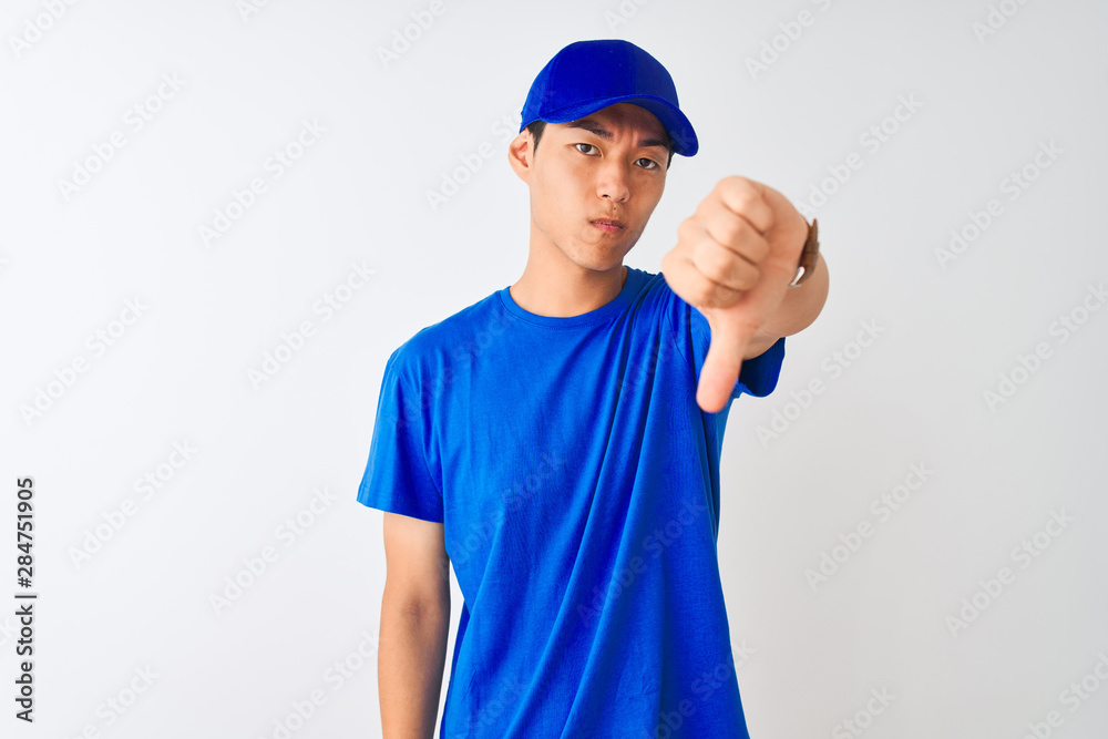 Chinese deliveryman wearing blue t-shirt and cap standing over isolated white background looking unhappy and angry showing rejection and negative with thumbs down gesture. Bad expression.