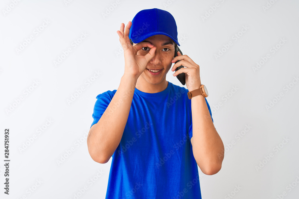 Chinese deliveryman wearing cap talking on the smartphone over isolated white background with happy face smiling doing ok sign with hand on eye looking through fingers Chinese deliveryman wearing cap talking on the smartphone over isolated white background with happy face smiling doing ok sign with hand on eye looking through fingers