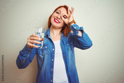 Young beautiful redhead woman drinking glass of water over white isolated background with happy face smiling doing ok sign with hand on eye looking through fingers