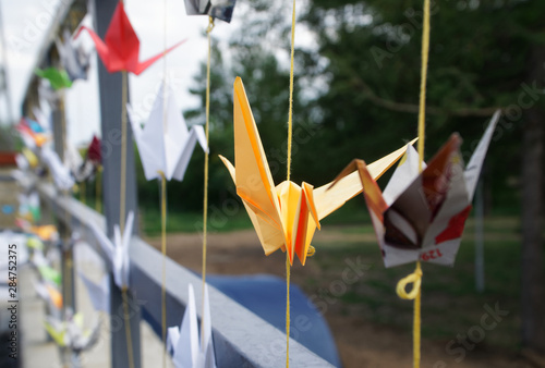Origami multicolored paper cranes tied to threads on the fence of a city park. Shallow depth of field
