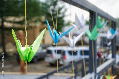 Origami multicolored paper cranes tied with thread to a fence near a parking lot. Shallow depth of field