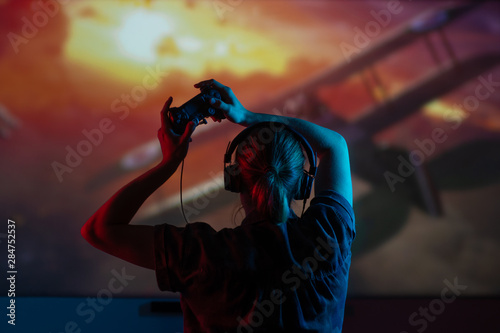 A girl gamer plays games with a gamepad, joystick on a large screen, with bright light and a dark room. eSports, gaming, streaming.bright background © Anton
