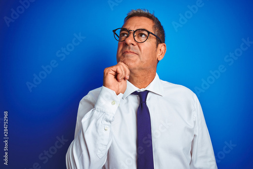 Handsome middle age businessman wearing glasses standing over isolated blue background serious face thinking about question, very confused idea