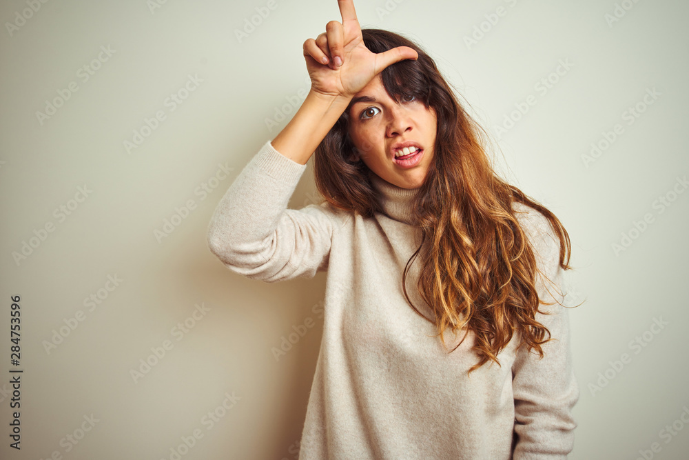 Fototapeta premium Young beautiful woman wearing winter sweater standing over white isolated background making fun of people with fingers on forehead doing loser gesture mocking and insulting.