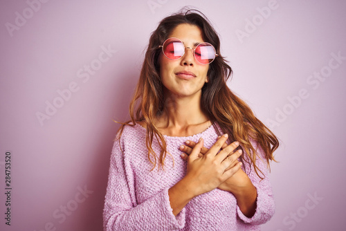 Young beautiful woman wearing fashion sunglasses standing over pink isolated background smiling with hands on chest with closed eyes and grateful gesture on face. Health concept.