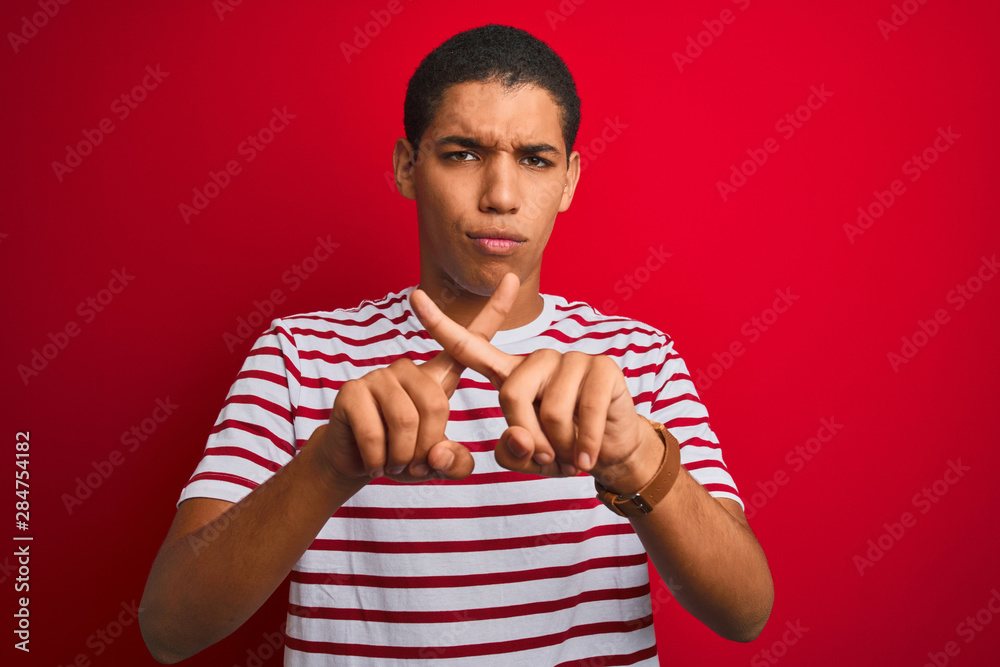 Young handsome arab man wearing striped t-shirt over isolated red background Rejection expression crossing fingers doing negative sign Young handsome arab man wearing striped t-shirt over isolated red background Rejection expression crossing fingers doing negative sign