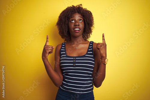 Young african afro woman wearing striped t-shirt over isolated yellow background amazed and surprised looking up and pointing with fingers and raised arms.