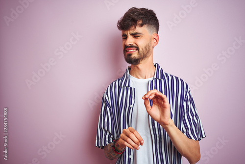 Young man with tattoo wearing striped shirt standing over isolated pink background disgusted expression, displeased and fearful doing disgust face because aversion reaction. With hands raised. 