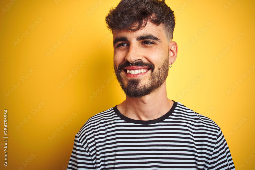 Young man with tattoo wearing striped t-shirt standing over isolated yellow background looking away to side with smile on face, natural expression. Laughing confident. Young man with tattoo wearing striped t-shirt standing over isolated yellow background looking away to side with smile on face, natural expression. Laughing confident.