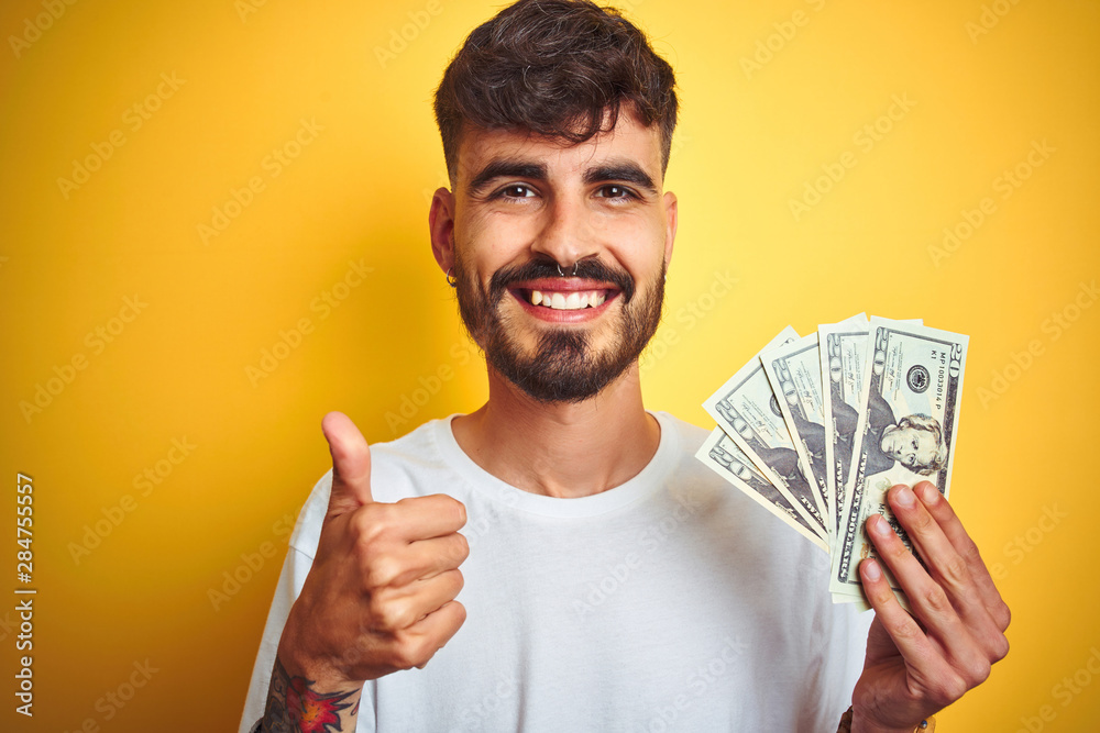 Young man with tattoo holding dollars standing over isolated yellow background happy with big smile doing ok sign, thumb up with fingers, excellent sign Young man with tattoo holding dollars standing over isolated yellow background happy with big smile doing ok sign, thumb up with fingers, excellent sign