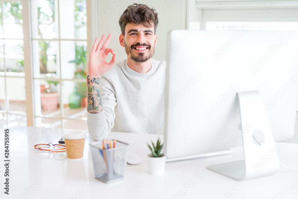 Young man working using computer doing ok sign with fingers, excellent symbol Young man working using computer doing ok sign with fingers, excellent symbol