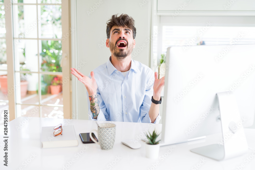 Young business man working using computer crazy and mad shouting and yelling with aggressive expression and arms raised. Frustration concept.
