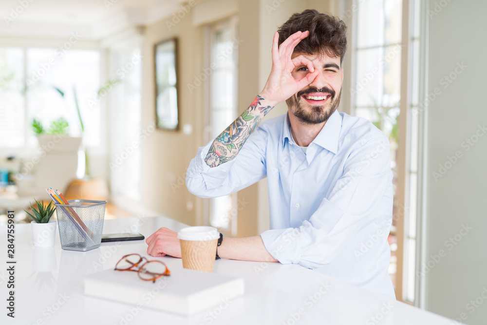 Young business man working doing ok gesture with hand smiling, eye looking through fingers with happy face.