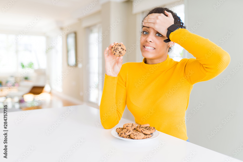Young african american girl eating chocolate chips cookies as sweet snack stressed with hand on head, shocked with shame and surprise face, angry and frustrated. Fear and upset for mistake.