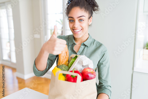 Young african american girl holding paper bag of groceries from supermarket happy with big smile doing ok sign  thumb up with fingers  excellent sign