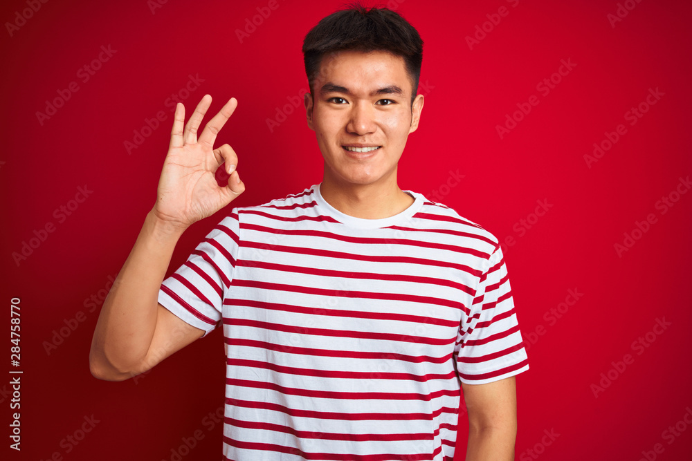 Young asian chinese man wearing striped t-shirt standing over isolated red background smiling positive doing ok sign with hand and fingers. Successful expression. Young asian chinese man wearing striped t-shirt standing over isolated red background smiling positive doing ok sign with hand and fingers. Successful expression.