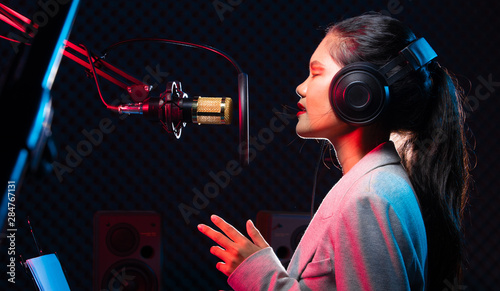 Fotografie Asian Teenager Woman black hair earmuff headphone sing a song loudly power sound over hanging microphone condenser, speakers, composer note book and monitor