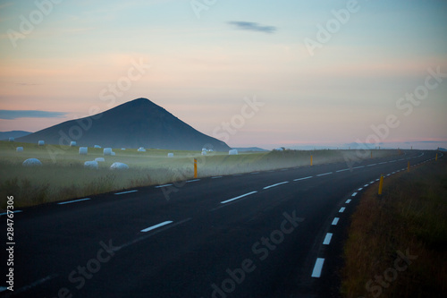 Summertime view of road near farm with hay bales in white plastic film stacked on a huge field in farm at Iceland.