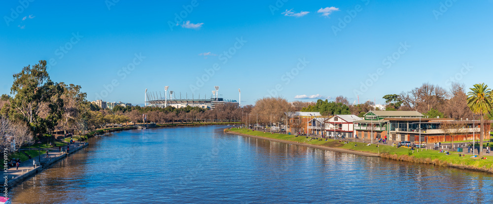 A large panorama of the Yarra River with the boathouses of Alexandra Gardens in the foreground and the Melbourne Cricket Ground in the distance