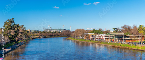 A large panorama of the Yarra River with the boathouses of Alexandra Gardens in the foreground and the Melbourne Cricket Ground in the distance