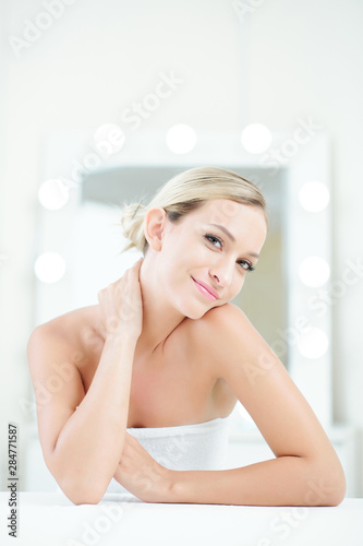 Positive beautiful young woman wrapped in towel after taking bath touching her soft skin and looking at camera