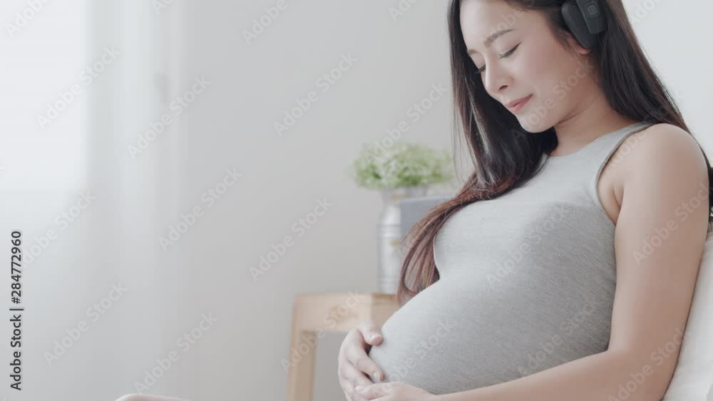 pregnant Asian mother open the music headphone and put hands her tummy with soft touch to make her child feeling relaxed on bedroom in the morning time pregnant Asian mother open the music headphone and put hands her tummy with soft touch to make her child feeling relaxed on bedroom in the morning time