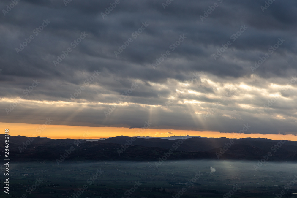 Sun rays coming down from some clouds over a valley filled by fog, illuminating part of it