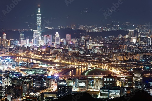 Night scenery of Downtown Taipei, the vibrant capital city of Taiwan, with view of landmark Tower among high-rise buildings in Xinyi Financial District & bridges over Keelung River in blue twilight