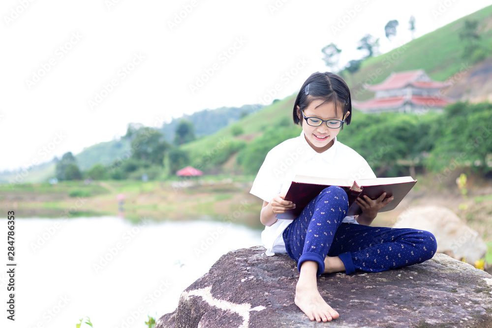 Cute asian little girl with glasses sitting near lake on stone reading a books