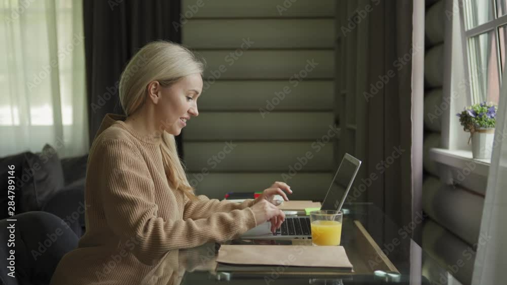 Medium shot of happy young woman typing on laptop computer sitting at desk at home and smiling cheerfully. Female freelancer enjoying networking, side view tracking right