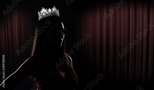 Portrait of Miss Pageant Beauty Contest in sequin Evening Ball Gown long dress with sparkle light Diamond Crown, silhouette low key exposure with curtain, studio lighting dark background dramatic